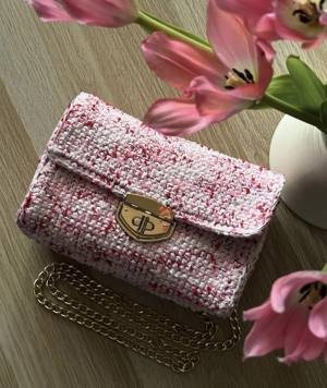 Handmade crochet flap bag in white yarn flecked with pink and red, with a gold clasp and gold chain strap, beside pink tulips