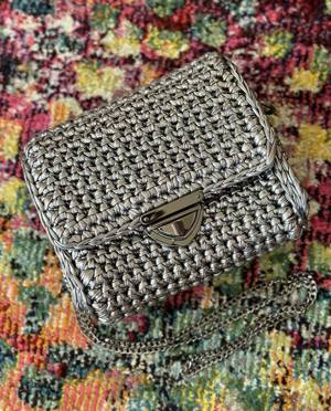 Handmade crochet box bag in silver metallic ribbon yarn with a black-nickel turn-lock clasp and chain strap, on a patterned rug